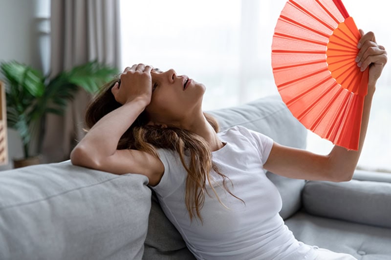 side view of a young woman, looking exhausted, using a paper fan to cool off in her apartment.