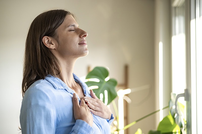A woman with brown hair, wearing a light blue button-down shirt, stands with her eyes closed and hands resting gently over her chest.